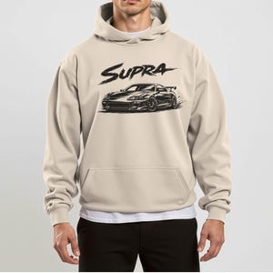 Könnte beinhalten: Beigefarbener Kapuzenpullover mit einer Vordertasche und einer Kapuze. Der Kapuzenpullover zeigt eine schwarze Grafik eines Autos und das Wort "Supra" in einer stilisierten Schriftart. Die Autografik ist eine detaillierte Illustration eines Sportwagens.