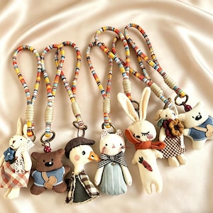 May include: A collection of handmade fabric keychains featuring various animal designs, including a bunny, bear, duck, cat, and star-shaped character. Each keychain has a colorful, patterned lanyard and is adorned with fabric clothing and accessories.