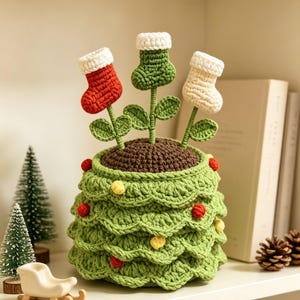 May include: A crocheted Christmas tree-shaped pot with three stems topped with small red, green, and white stockings. The green pot features scalloped layers decorated with small red and yellow pom-poms, and the top is filled with brown yarn.