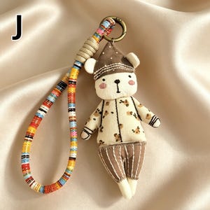 May include: A handmade bear keychain with a colorful strap. The bear is wearing a brown hat and a cream-colored outfit with brown floral patterns. The keychain has a metal ring and a braided strap with various colors.