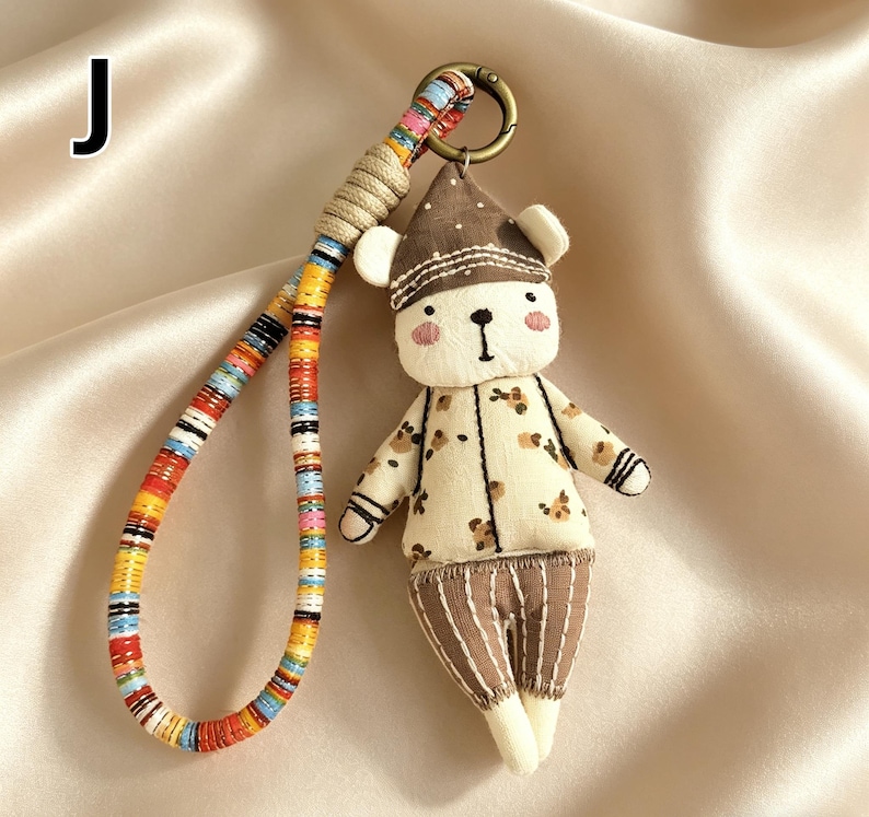 May include: A handmade bear keychain with a colorful strap. The bear is wearing a brown hat and a cream-colored outfit with brown floral patterns. The keychain has a metal ring and a braided strap with various colors.