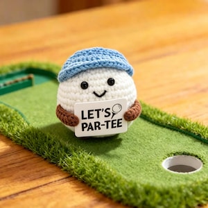 May include: A crocheted golf ball character wearing a blue hat and brown arms, holding a sign that reads "LET'S PAR-TEE." The character is on a miniature golf green with a hole and a wooden background.