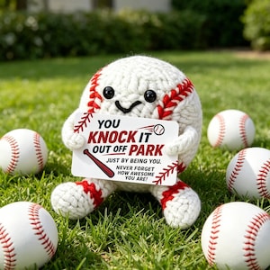 Puede incluir: Un personaje de béisbol tejido con una cara sonriente, sosteniendo un letrero que dice "You Knock It Out Off Park". El letrero también dice "Just by being you. Never forget how awesome you are!" Varias pelotas de béisbol están esparcidas en la hierba.