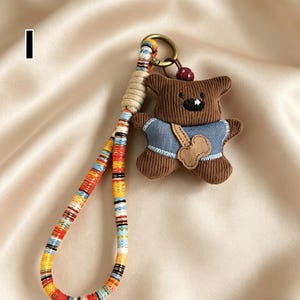 May include: A brown teddy bear keychain with a blue shirt and a bone-shaped patch. The keychain has a colorful woven strap and a gold-colored ring. The bear has a black stitched mouth and a small red bead on top of its head.