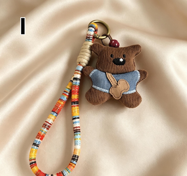 May include: A brown teddy bear keychain with a blue shirt and a bone-shaped patch. The keychain has a colorful woven strap and a gold-colored ring. The bear has a black stitched mouth and a small red bead on top of its head.