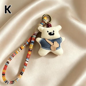 May include: A cream-colored bear-shaped keychain with a denim vest and a wooden-colored guitar. The keychain has a colorful woven strap and a gold-colored metal ring. The letter "K" is in the upper left corner.