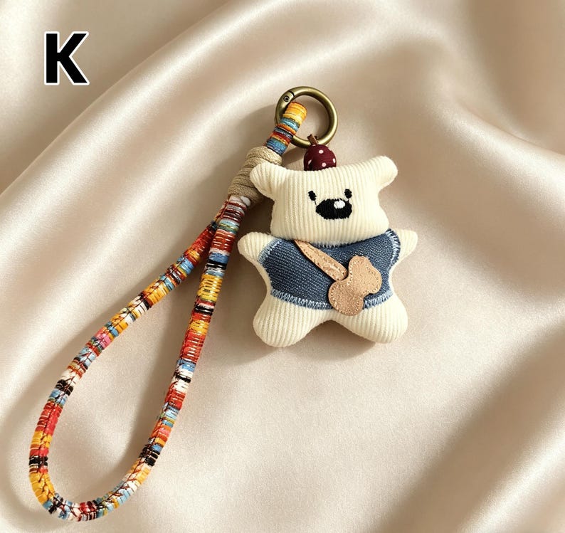 May include: A cream-colored bear-shaped keychain with a denim vest and a wooden-colored guitar. The keychain has a colorful woven strap and a gold-colored metal ring. The letter "K" is in the upper left corner.