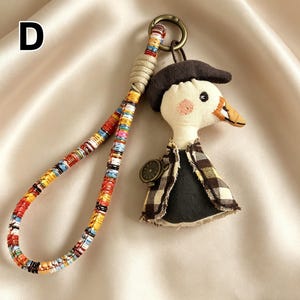 May include: A whimsical keychain featuring a plush duck character wearing a brown hat and plaid vest. The keychain includes a colorful woven strap and a small, decorative watch. The letter "D" is in the upper left corner.