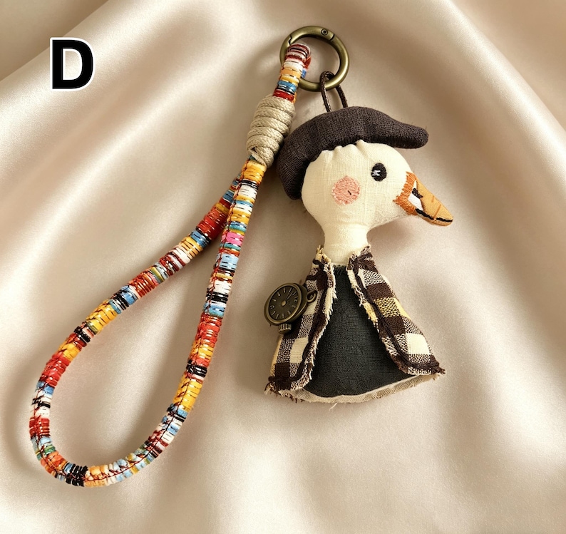 May include: A whimsical keychain featuring a plush duck character wearing a brown hat and plaid vest. The keychain includes a colorful woven strap and a small, decorative watch. The letter "D" is in the upper left corner.
