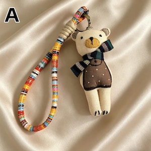 May include: A beige teddy bear keychain with a brown vest and a striped scarf. The keychain has a colorful, patterned strap and a metal clasp. The bear's features are embroidered with black and yellow thread.