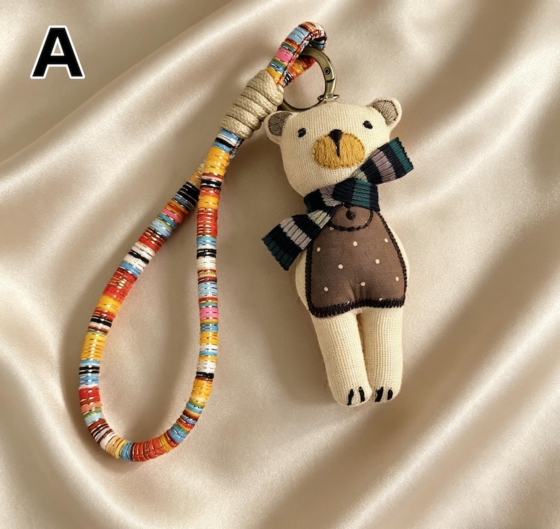 May include: A beige teddy bear keychain with a brown vest and a striped scarf. The keychain has a colorful, patterned strap and a metal clasp. The bear's features are embroidered with black and yellow thread.