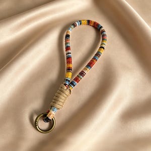 May include: A colorful keychain wrist strap with a woven design. The strap features a loop of tightly wound, multi-colored threads in red, yellow, blue, and black. A tan woven section connects the loop to a gold-colored metal ring.