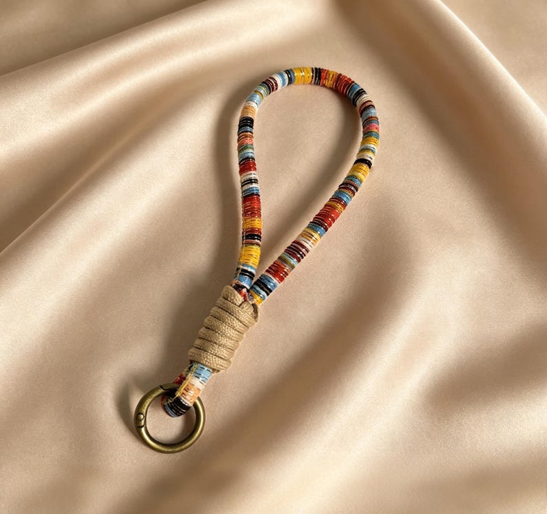 May include: A colorful keychain wrist strap with a woven design. The strap features a loop of tightly wound, multi-colored threads in red, yellow, blue, and black. A tan woven section connects the loop to a gold-colored metal ring.