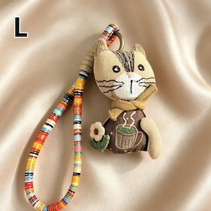 May include: A beige cat-shaped keychain with a brown and white striped head, wearing a brown vest with a coffee cup design. The keychain has a colorful, patterned strap and a metal ring.