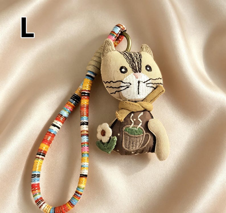 May include: A beige cat-shaped keychain with a brown and white striped head, wearing a brown vest with a coffee cup design. The keychain has a colorful, patterned strap and a metal ring.