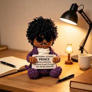 May include: A crocheted doll with dark curly hair, black sunglasses, and a purple jacket, holding a white sign that reads: EMOTIONAL SUPPORT PRINCE Dearly beloved, I'm here to get through this thing called life. The doll has a white ruffled collar.