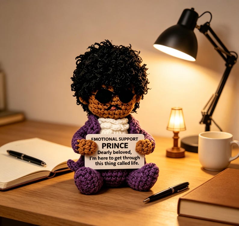 May include: A crocheted doll with dark curly hair, black sunglasses, and a purple jacket, holding a white sign that reads: EMOTIONAL SUPPORT PRINCE Dearly beloved, I'm here to get through this thing called life. The doll has a white ruffled collar.