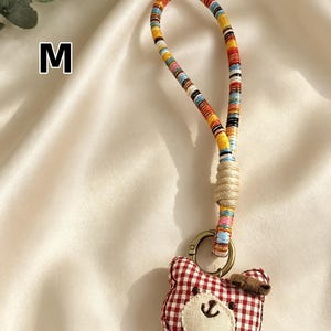 May include: A handmade keychain featuring a red and white gingham bear charm with a stitched face and a small bow. The keychain has a colorful, striped fabric strap and a metal clasp. The letter "M" is visible in the background.