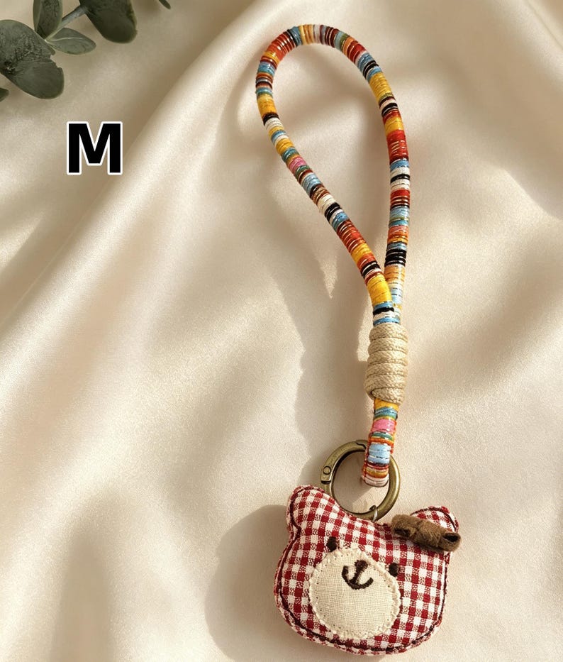 May include: A handmade keychain featuring a red and white gingham bear charm with a stitched face and a small bow. The keychain has a colorful, striped fabric strap and a metal clasp. The letter "M" is visible in the background.