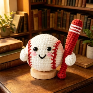 Puede incluir: Una pelota de béisbol de ganchillo con una cara sonriente, mejillas rosadas y costuras rojas, sosteniendo un bate a rayas rojas y blancas. La pelota de béisbol está sobre un soporte de madera. El fondo incluye una estantería y libros.