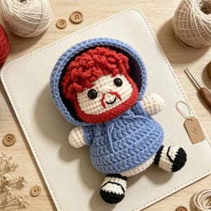 Könnte beinhalten: Eine handgefertigte Häkelpuppe mit lockigem rotem Haar, rotem Bart und schwarzen Sicherheitsaugen. Die Puppe trägt einen hellblauen Kapuzenpullover mit Kordelzug sowie schwarz-weiße Schuhe. Die Figur ist aus Garn gefertigt.
