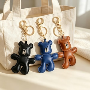 May include: Three bear-shaped keychains in black, blue, and brown, each featuring a gold-toned metal lobster clasp and split ring. The bears have stitched facial details and a smooth finish, hanging from a light-coloured canvas tote bag.