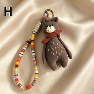 May include: A brown bear keychain with a red scarf and a colorful strap. The bear has white accents on its face and small stitched details on its body. The strap is made of colorful beads and rope.