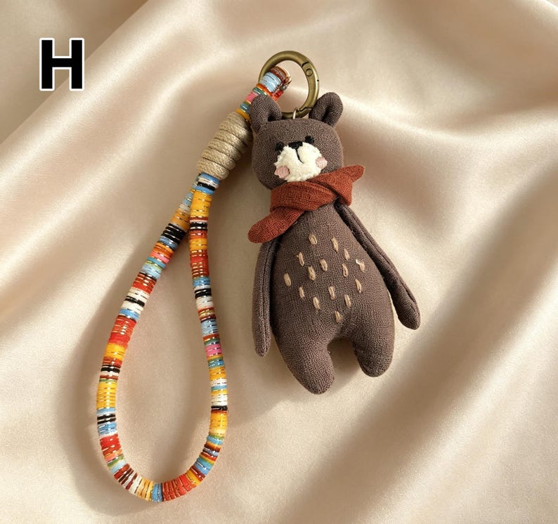 May include: A brown bear keychain with a red scarf and a colorful strap. The bear has white accents on its face and small stitched details on its body. The strap is made of colorful beads and rope.