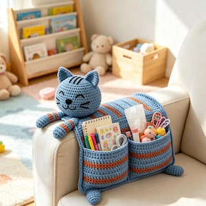 以下が含まれることがあります： アームレストに掛けられた青とオレンジの縞模様の猫型かぎ針編みオーガナイザー。猫の頭部には刺繍が施され、胴体部分にはノート、ペン、ティッシュ、小さなおもちゃが入った2つの深いポケットが付いています。