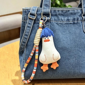 Könnte beinhalten: Eine Jeans-Tragetasche mit einem bunten Riemen und einem niedlichen Enten-Schlüsselanhänger. Der Schlüsselanhänger ist weiß mit blauen Haaren und orangefarbenen Füßen. Die Tasche hat einen Schultergurt und Metallbeschläge.
