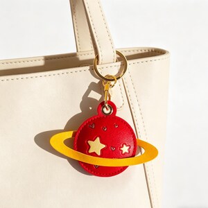 May include: A red planet-shaped keychain with a yellow ring, gold-colored hardware, and a small star and heart design. The keychain is attached to a cream-colored tote bag.