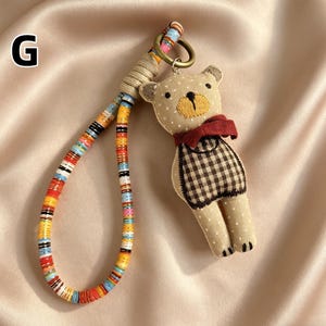 May include: A beige teddy bear keychain with a red scarf and a checkered pattern vest. The keychain has a colorful beaded strap and a metal ring. The bear is made of soft fabric and has embroidered details.