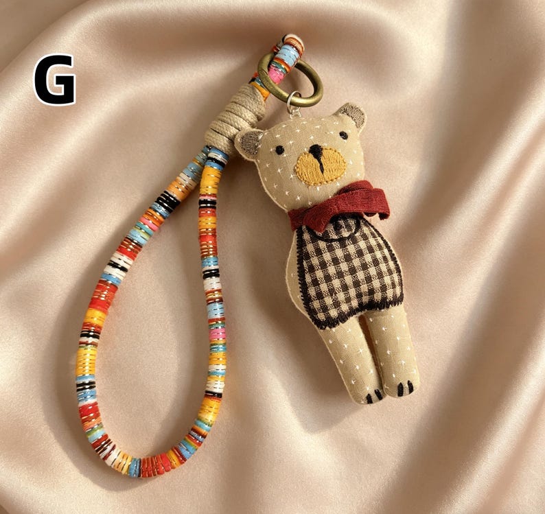 May include: A beige teddy bear keychain with a red scarf and a checkered pattern vest. The keychain has a colorful beaded strap and a metal ring. The bear is made of soft fabric and has embroidered details.