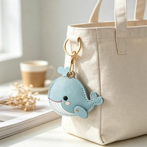May include: A light blue whale-shaped keychain made of smooth material with stitched edges, a small black eye, a pink cheek detail, and white line accents on the belly, attached to a gold-toned metal ring and clasp hanging from a cream canvas tote bag.