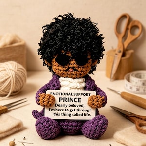 May include: A crocheted doll with dark curly hair, black sunglasses, and a purple suit, holding a white sign that reads: EMOTIONAL SUPPORT PRINCE. Dearly beloved, I'm here to get through this thing called life. The doll has a white ruffled collar.