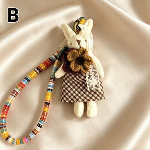 May include: A handmade bunny doll with a brown and white checkered dress and a large flower accent. The doll has a colorful beaded strap. The letter "B" is in the upper left corner.