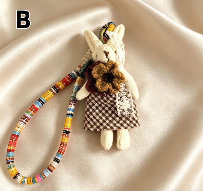 May include: A handmade bunny doll with a brown and white checkered dress and a large flower accent. The doll has a colorful beaded strap. The letter "B" is in the upper left corner.
