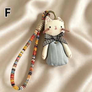 May include: A handmade cat-shaped keychain with a white face, pink accents, and a black and white striped bow tie. The cat wears a light blue dress and has a colorful patterned strap. A gold-colored ring is attached to the top.