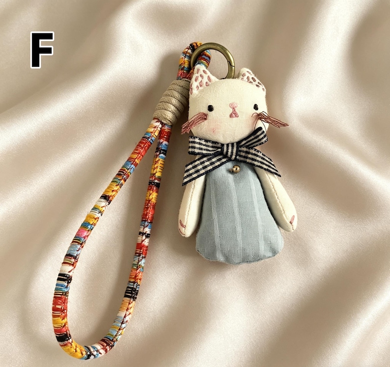 May include: A handmade cat-shaped keychain with a white face, pink accents, and a black and white striped bow tie. The cat wears a light blue dress and has a colorful patterned strap. A gold-colored ring is attached to the top.
