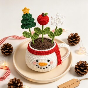 May include: A white crocheted mug shaped like a snowman with a red scarf, containing three crocheted stems topped with a green Christmas tree, a red apple, and a white snowflake. The mug features black eyes, a carrot nose, and pink cheeks.