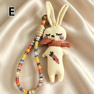 May include: A cream-colored plush bunny keychain with long ears, closed eyes, and a rust-colored scarf. The bunny has a carrot embroidered on its body. The keychain strap is multi-colored with a woven beige section.
