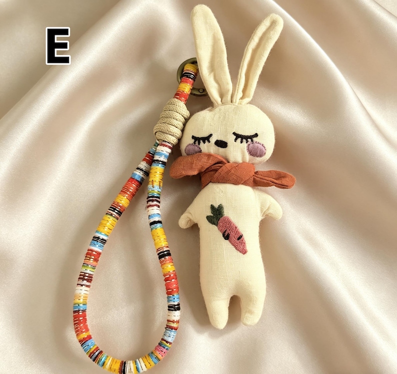 May include: A cream-colored plush bunny keychain with long ears, closed eyes, and a rust-colored scarf. The bunny has a carrot embroidered on its body. The keychain strap is multi-colored with a woven beige section.