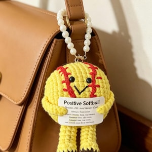 Peut inclure: Un porte-clés en forme de balle de softball jaune crochetée, avec des coutures rouges et un visage souriant, tenant une pancarte blanche avec l'inscription "Positive Softball". Le porte-clés est attaché à une chaîne de perles, suspendu à un sac à main marron.