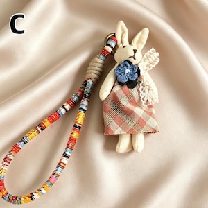 May include: A handmade bunny keychain with a plaid dress and a blue flower. The keychain has a colorful woven strap and a beige cord loop. The bunny has cream-colored fur and lace wings.