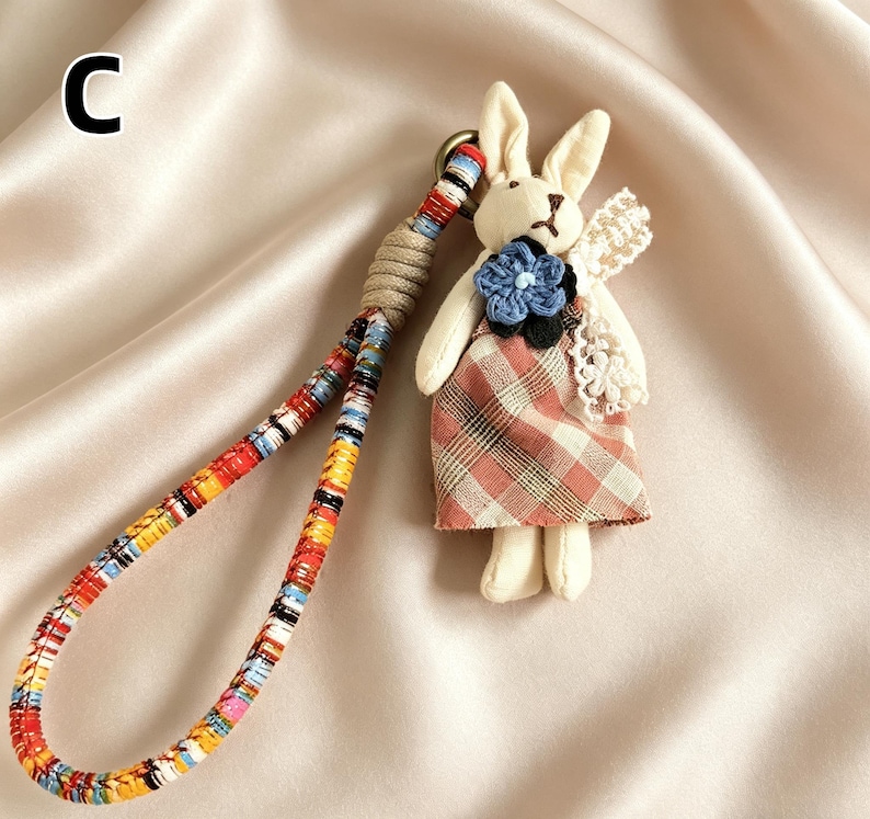 May include: A handmade bunny keychain with a plaid dress and a blue flower. The keychain has a colorful woven strap and a beige cord loop. The bunny has cream-colored fur and lace wings.