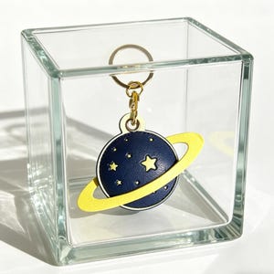 May include: A navy blue planet-shaped keychain with a yellow ring and gold-colored hardware. The planet has a constellation of small white stars and a large yellow star. The keychain is displayed inside a clear glass cube.