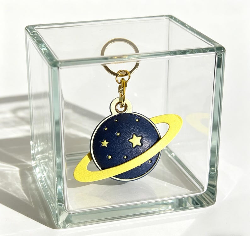 May include: A navy blue planet-shaped keychain with a yellow ring and gold-colored hardware. The planet has a constellation of small white stars and a large yellow star. The keychain is displayed inside a clear glass cube.