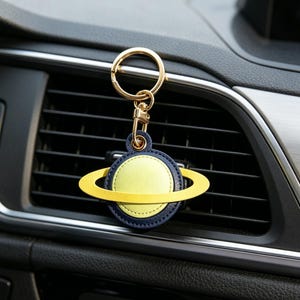 May include: A planet-shaped keychain with a gold ring and clasp. The planet design features a yellow center, a navy blue border, and a yellow ring around it. The keychain is hanging in front of a car's air vent.