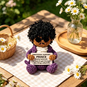 May include: A crocheted doll with dark curly hair, black sunglasses, and a purple suit, holding a white sign that reads: EMOTIONAL SUPPORT PRINCE Dearly beloved, I'm here to get through this thing called life. The doll has a tan complexion.