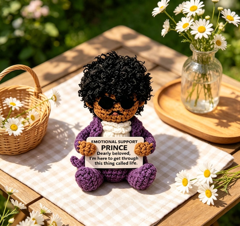 May include: A crocheted doll with dark curly hair, black sunglasses, and a purple suit, holding a white sign that reads: EMOTIONAL SUPPORT PRINCE Dearly beloved, I'm here to get through this thing called life. The doll has a tan complexion.